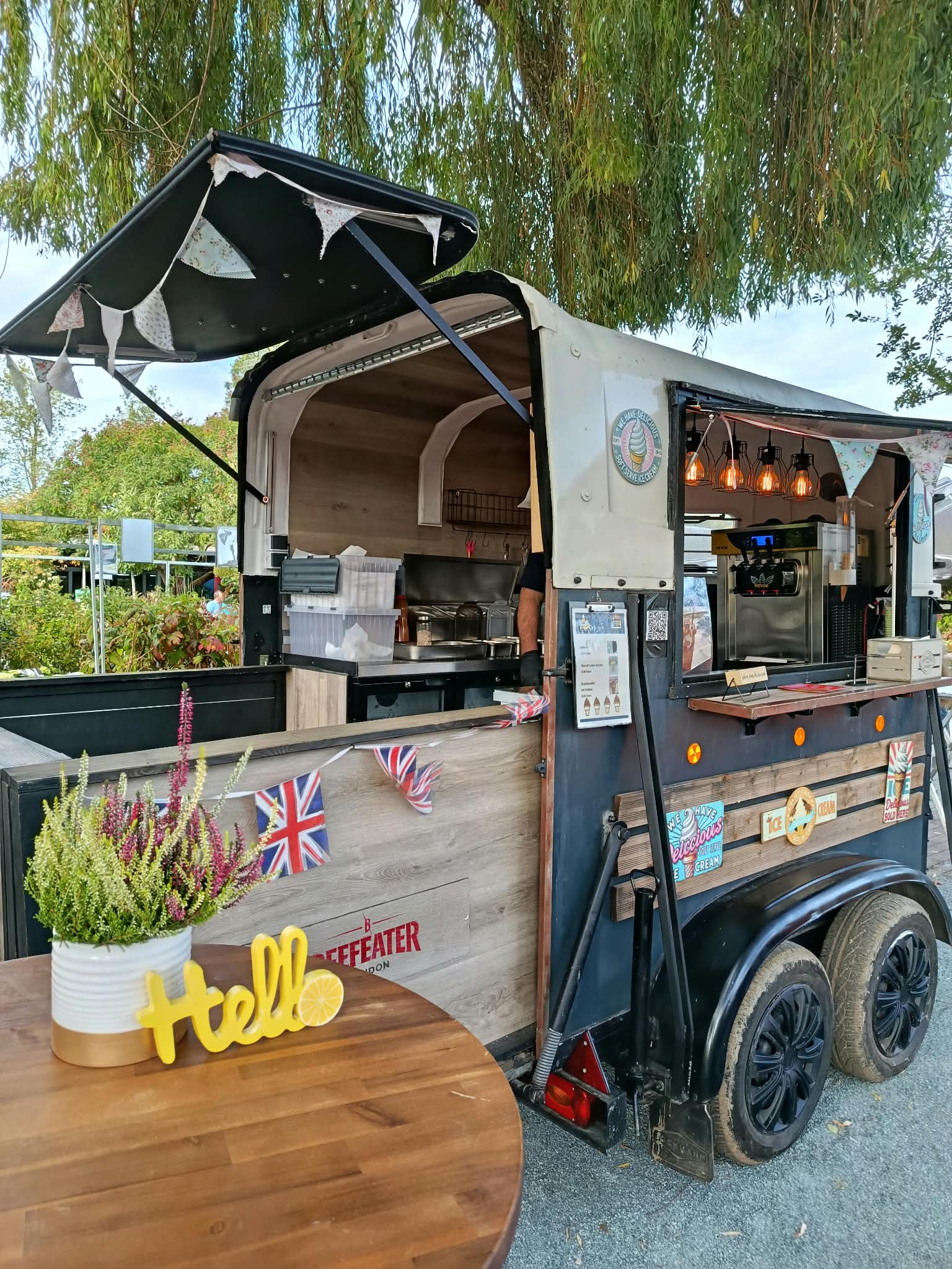 Unser Foodtrailer unter einer Weide, mit Holztheke, Wimpeln und warmen Pendelleuchten